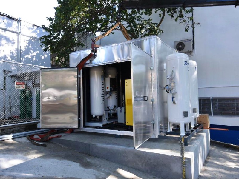 Foto da miniusina de oxigênio, instalada na área externa ao PS. O contâiner metálico está posicionado na frente de uma árvore sob uma base de concreto. As portas estão abertas e dentro estão os cilindros e equipamentos para geração do insumo. Fora do contâiner também há dois grandes cilindros